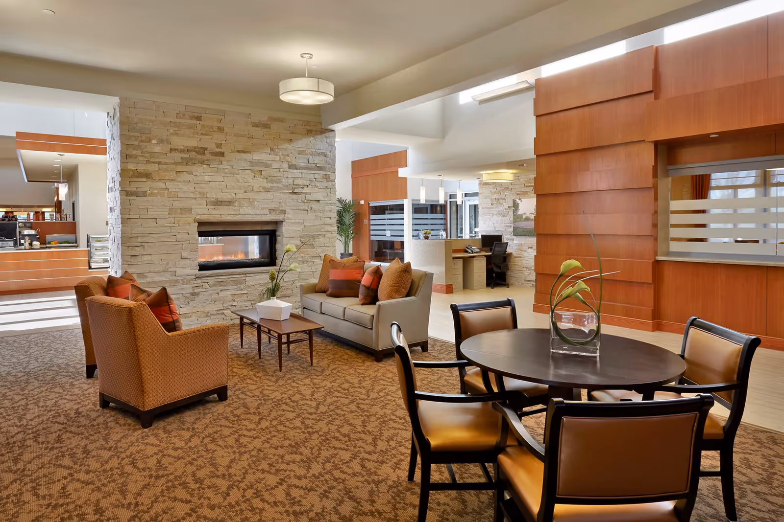 A spacious and well-lit common area in a senior living facility featuring a stone fireplace, a beige sofa with orange and brown cushions, two matching armchairs, a wooden coffee table with a white vase, and a round dining table with four chairs. The room has carpeted flooring, wooden panel walls, and a reception desk in the background.