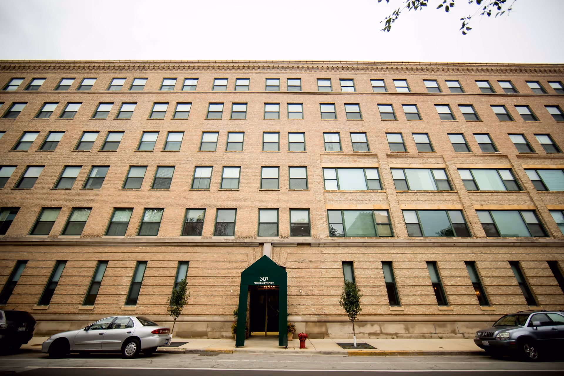 Exterior front view of a multi-story brick building with many windows, a green entrance canopy labeled 2437, two small trees on the sidewalk, and parked cars along the street.