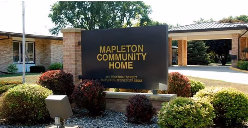 Sign reading "Mapleton Community Home" surrounded by shrubs in front of the facility entrance and brick building.