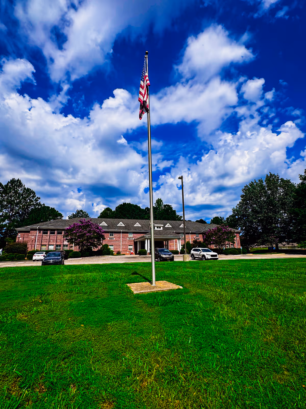 A large green lawn with an American flag on a flagpole in the center foreground. Behind the lawn is a two-story brick building with a covered entrance, several cars parked in front, and trees on either side. The sky is partly cloudy with patches of blue.