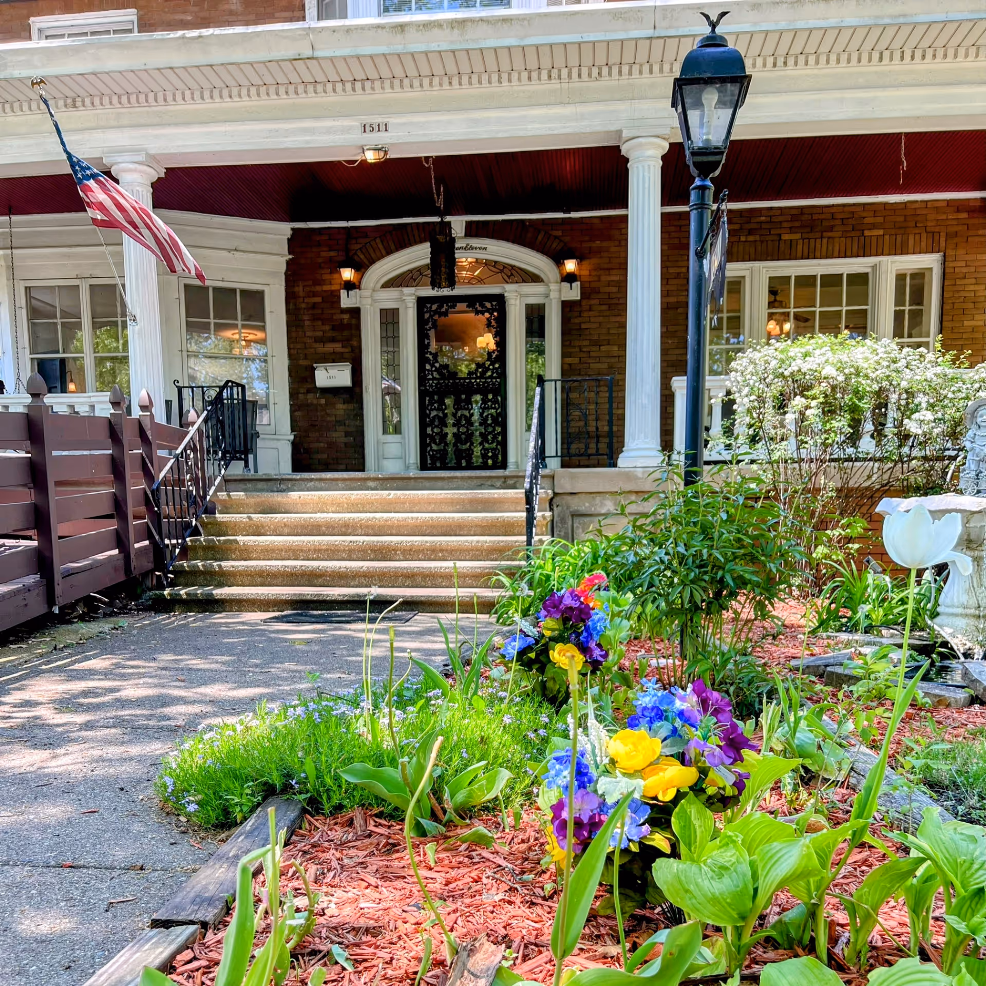 Front entrance of a senior living facility with a brick exterior, white columns, and a porch. There are steps leading up to a decorative black door with glass panels. An American flag is mounted on the left side of the porch. In the foreground, there is a garden bed with colorful flowers and green plants, along with a black lamppost and a white birdbath or fountain on the right side.