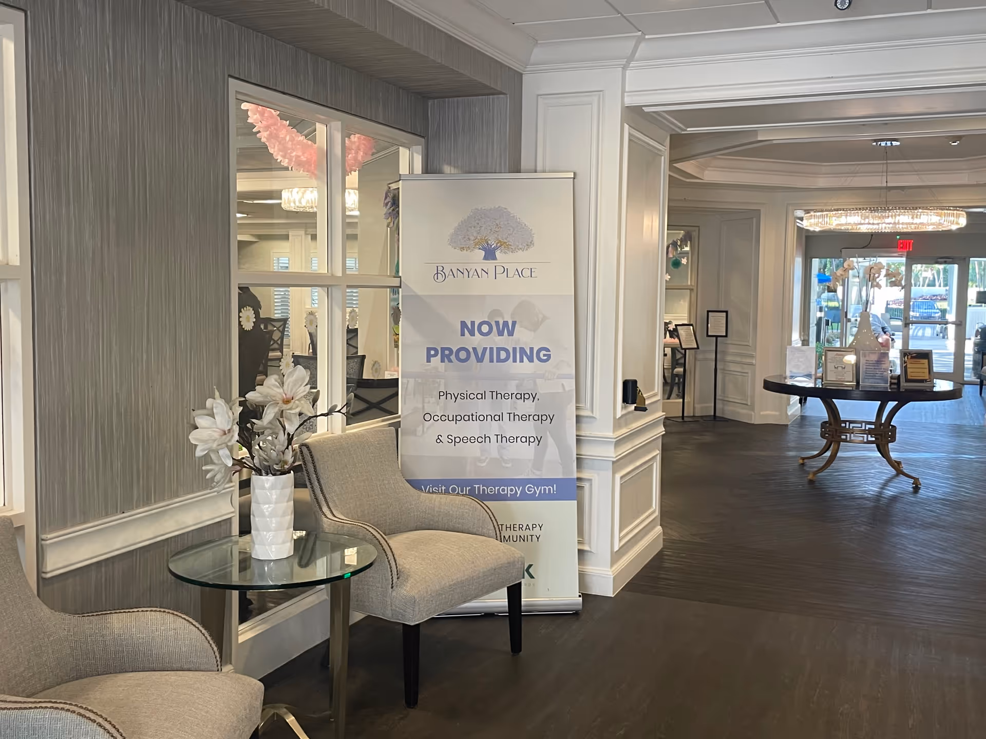 Well-lit lobby seating area with chairs, a side table with flowers, and a Banyan Place sign announcing therapy services.