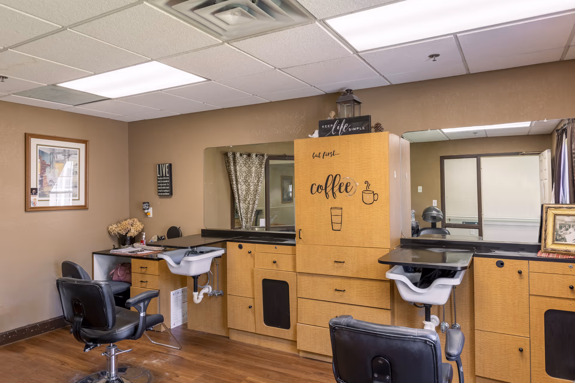 Interior view of a salon area within a senior living facility featuring two black salon chairs in front of wooden cabinetry with sinks, a large mirror, and wall decor including a sign that says 'but first... coffee'.