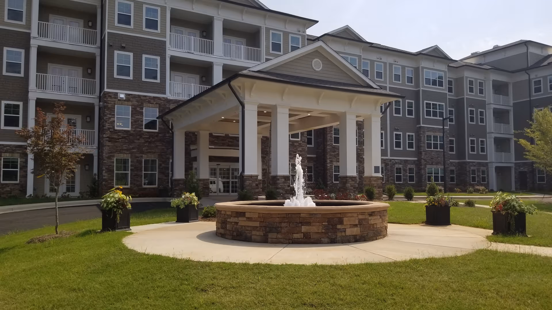 Front exterior view of a multi-story senior living facility with a covered entrance supported by white columns and a circular stone fountain in front. The building features a combination of stone and siding with multiple windows and balconies. There is a well-maintained lawn and several planters with greenery around the entrance area.