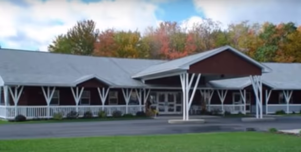 Front exterior of a single-story building with a covered entrance, white porch supports and autumn trees behind it.