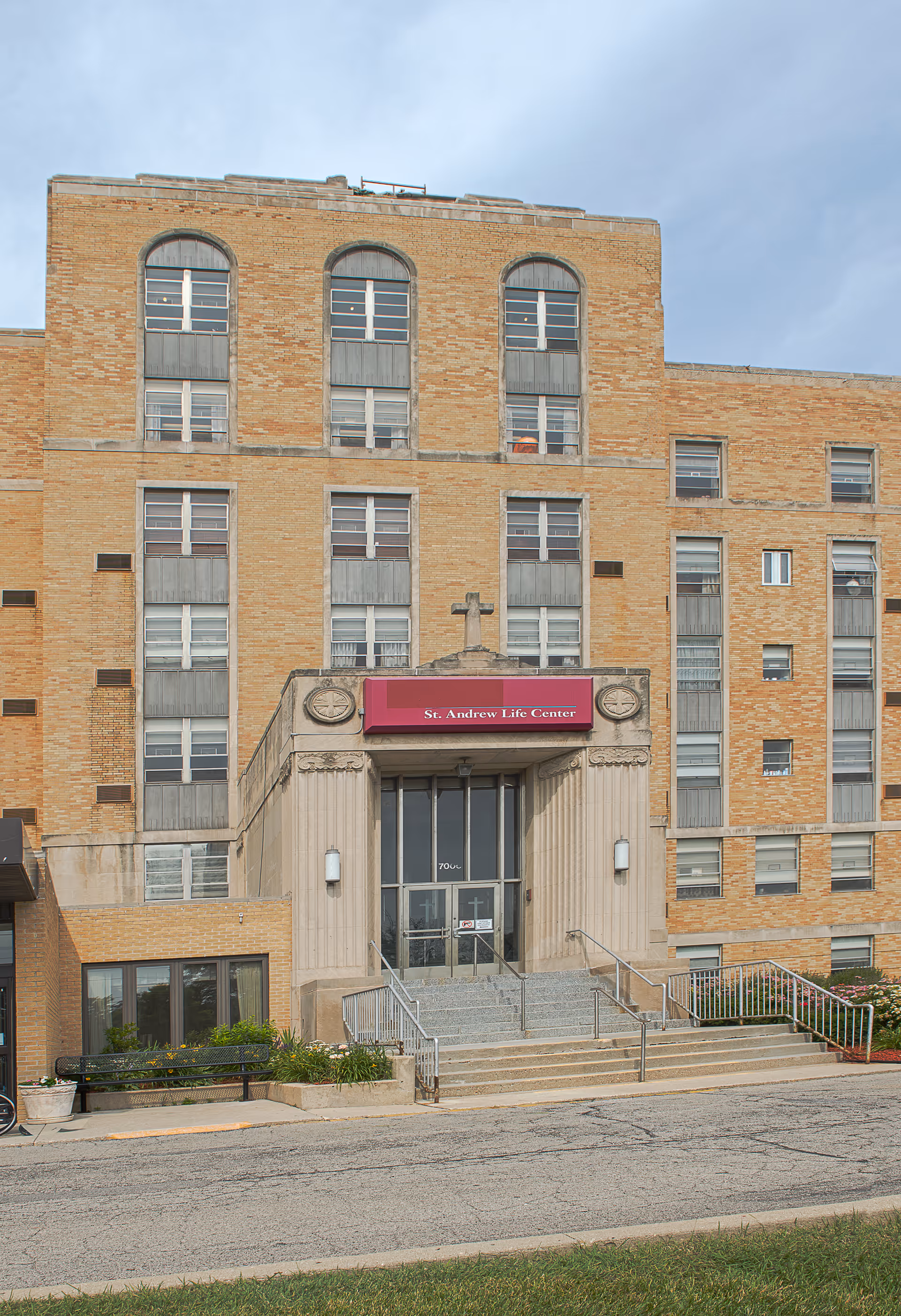 Front exterior view of a multi-story brick building with a central entrance labeled 'St. Andrew Life Center'. The entrance has steps leading up to glass doors, flanked by columns and outdoor lights. There is a small garden area and a bench near the entrance.