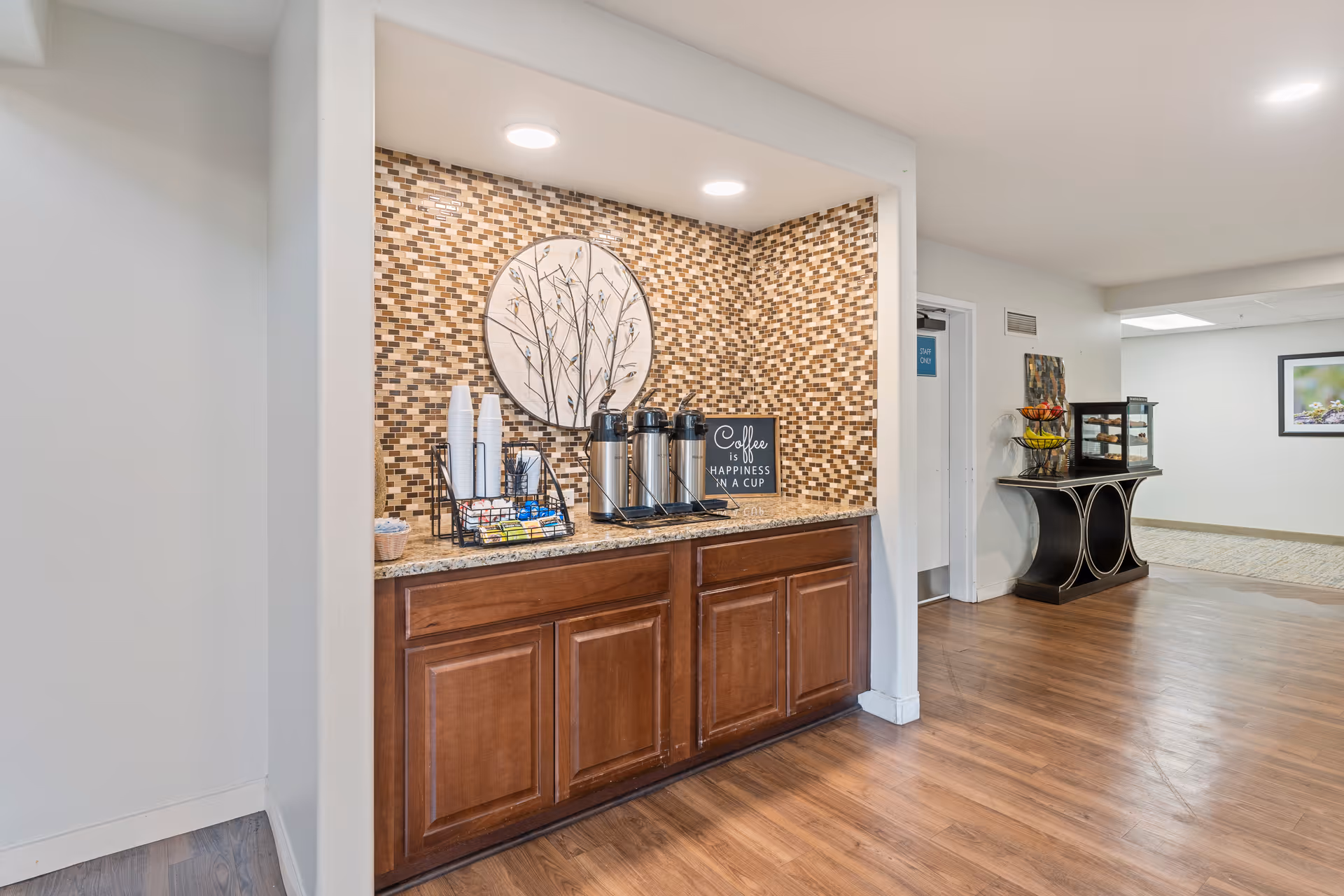 A coffee station in a senior living facility with a mosaic tile backsplash, three coffee dispensers, disposable cups, and a sign that reads 'Coffee is happiness in a cup.' The area has wooden cabinets below the countertop and recessed lighting above. In the background, there is a hallway with a table holding a fruit basket and a display case.