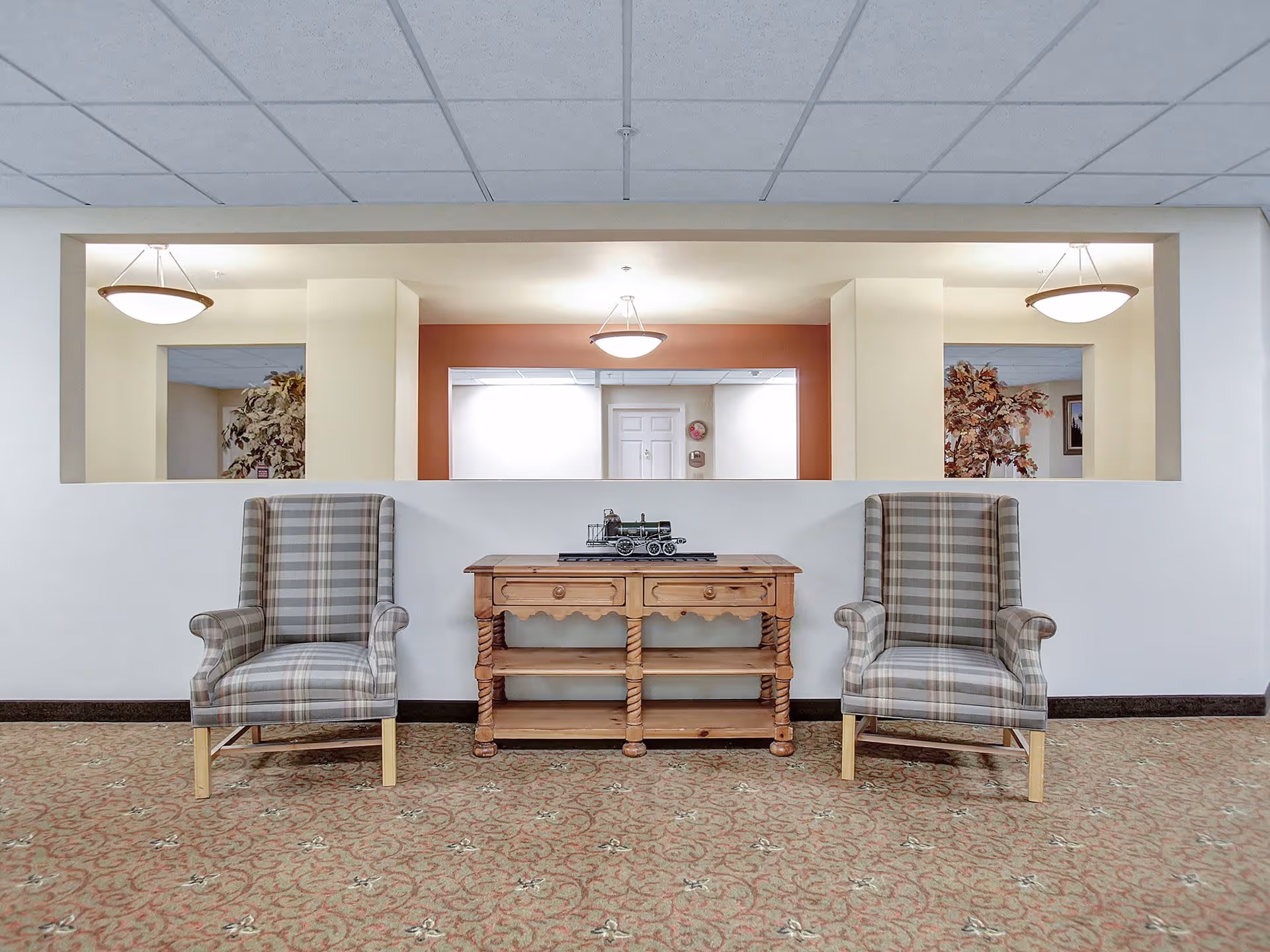 Two plaid upholstered armchairs flank a wooden console table with a decorative model train on top, set against a half wall with a large rectangular opening revealing a hallway with ceiling lights and plants in the background.