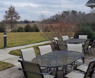Outdoor patio with metal tables and cushioned chairs overlooking a grassy lawn and trees under a cloudy sky.