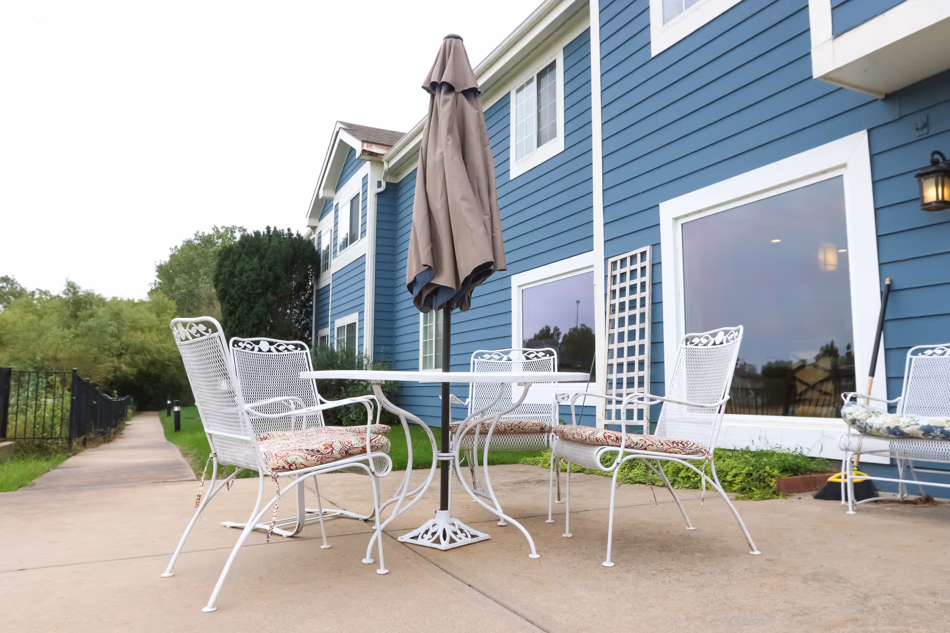 Outdoor patio area with a white metal table and four matching chairs with patterned cushions, situated next to a blue building with large windows. A closed beige umbrella is positioned in the center of the table. There is a paved walkway and greenery in the background.