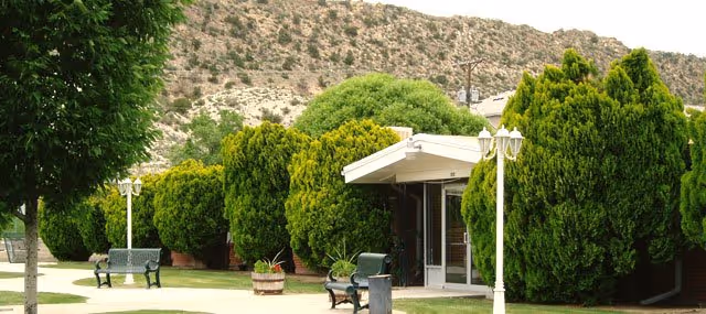 Outdoor area of Skyline Ridge Nursing and Rehabilitation Center featuring a pathway lined with green bushes and trees, benches, lamp posts, and a building entrance with a white awning. In the background, there is a rocky hill under a clear sky.