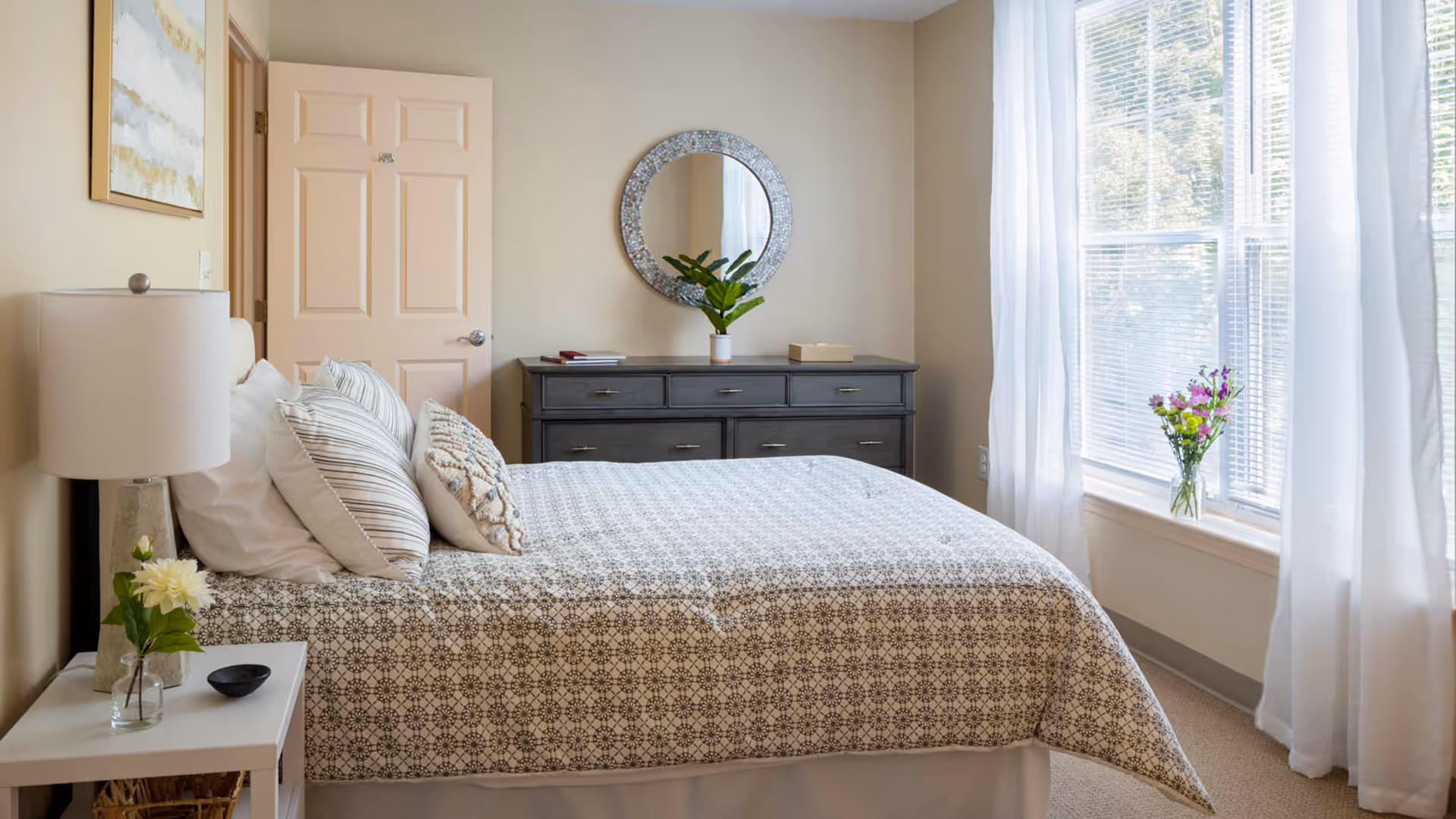 Sunlit bedroom with a patterned bed, bedside table and lamp, dresser with a round mirror, and a window with sheer curtains.
