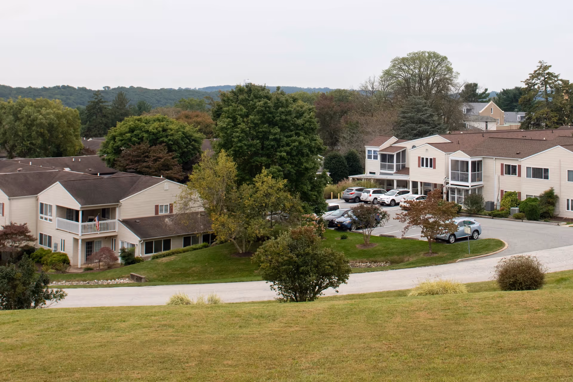 View of Glen Meadows Retirement Community showing multiple beige two-story residential buildings with brown roofs, surrounded by green trees and a grassy lawn. Several cars are parked in a parking lot near the buildings, and hills are visible in the background under an overcast sky.