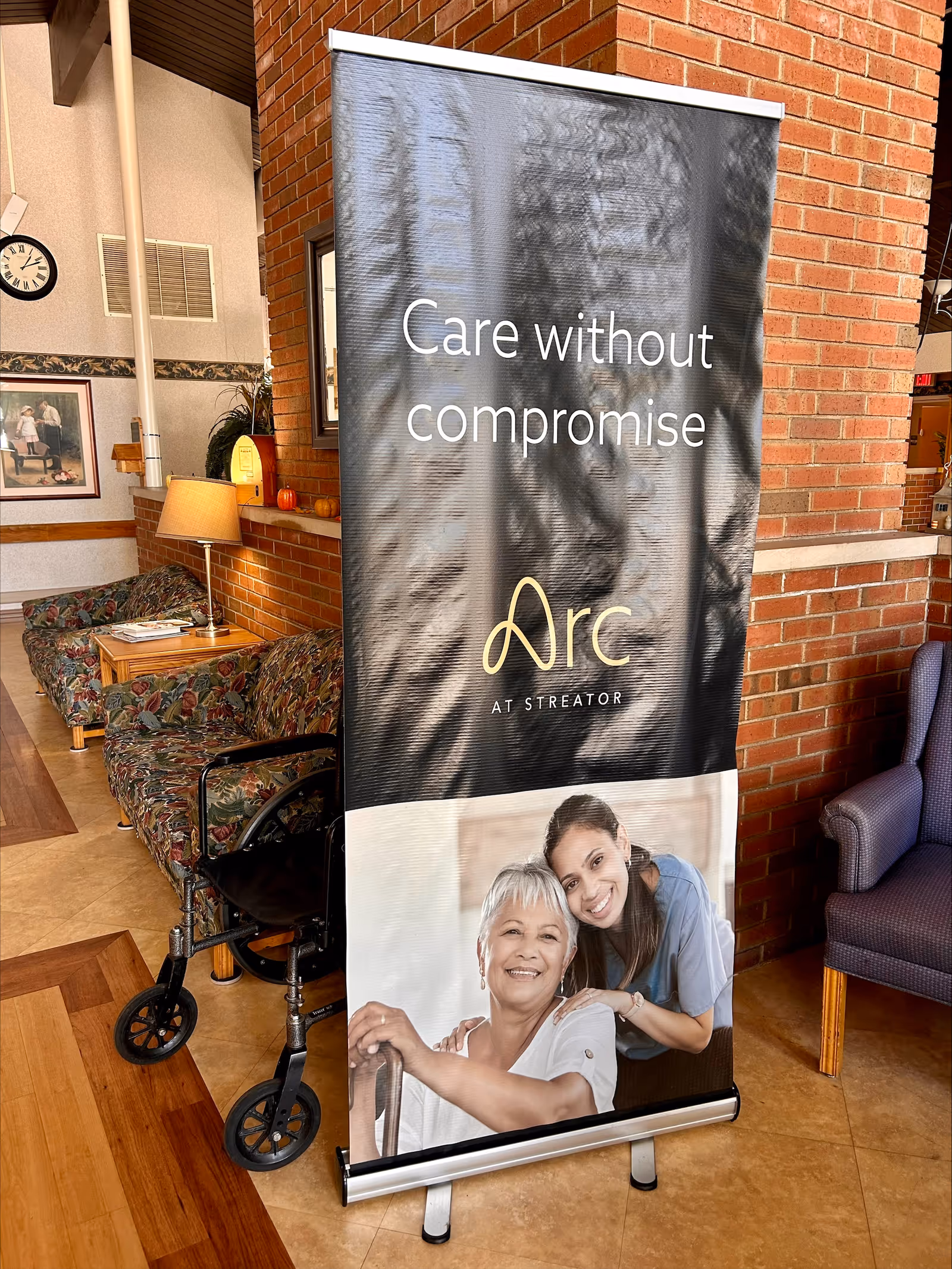 A standing promotional banner in a senior living lobby reading "Care without compromise" with chairs, a walker, and brick walls visible around it.