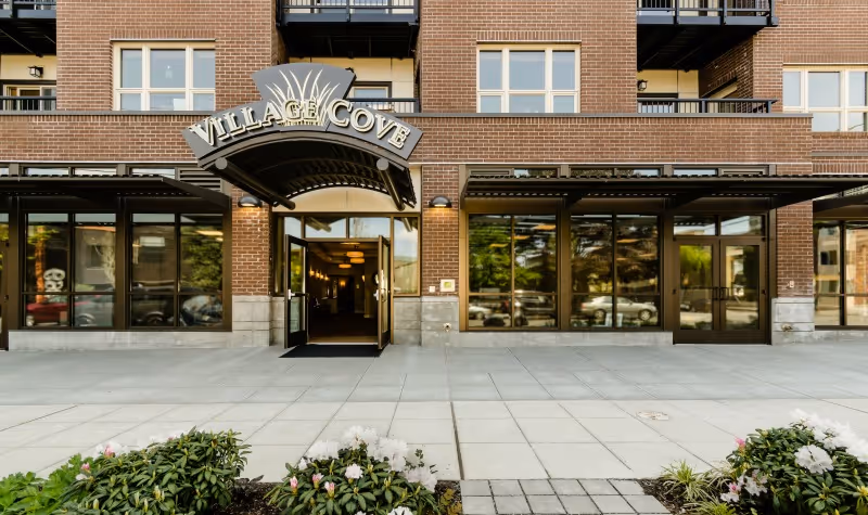 Entrance of a brick senior housing building with a curved awning reading "Village Cove" above glass doors and landscaped planters in front.