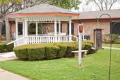 A white gazebo with a red roof surrounded by neatly trimmed bushes and green grass in front of a brick building, with a stone bench and a bird feeder visible on the lawn.