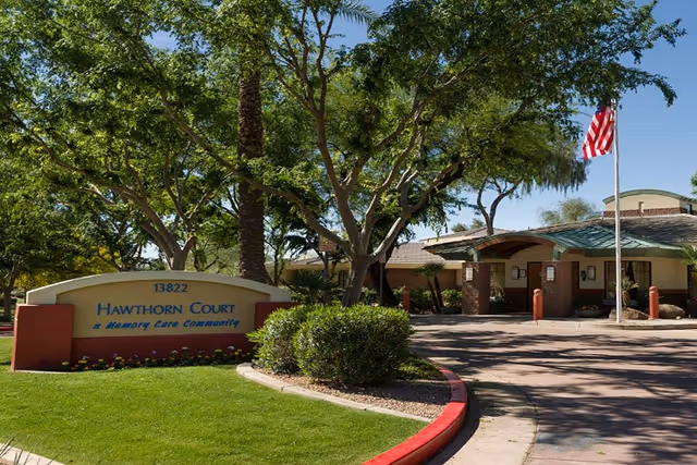 Exterior view of Hawthorn Court at Ahwatukee Memory Care facility with a sign displaying the name and address, surrounded by trees and landscaping, and an American flag on a flagpole near the entrance.