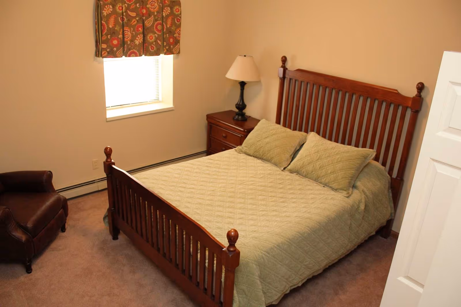 A bedroom with a wooden bed frame and matching headboard and footboard, covered with a light green quilt and two matching pillows. There is a wooden nightstand with a lamp on it next to the bed, a window with a floral valance, and a brown leather armchair in the corner. The walls are beige and the carpet is light brown.