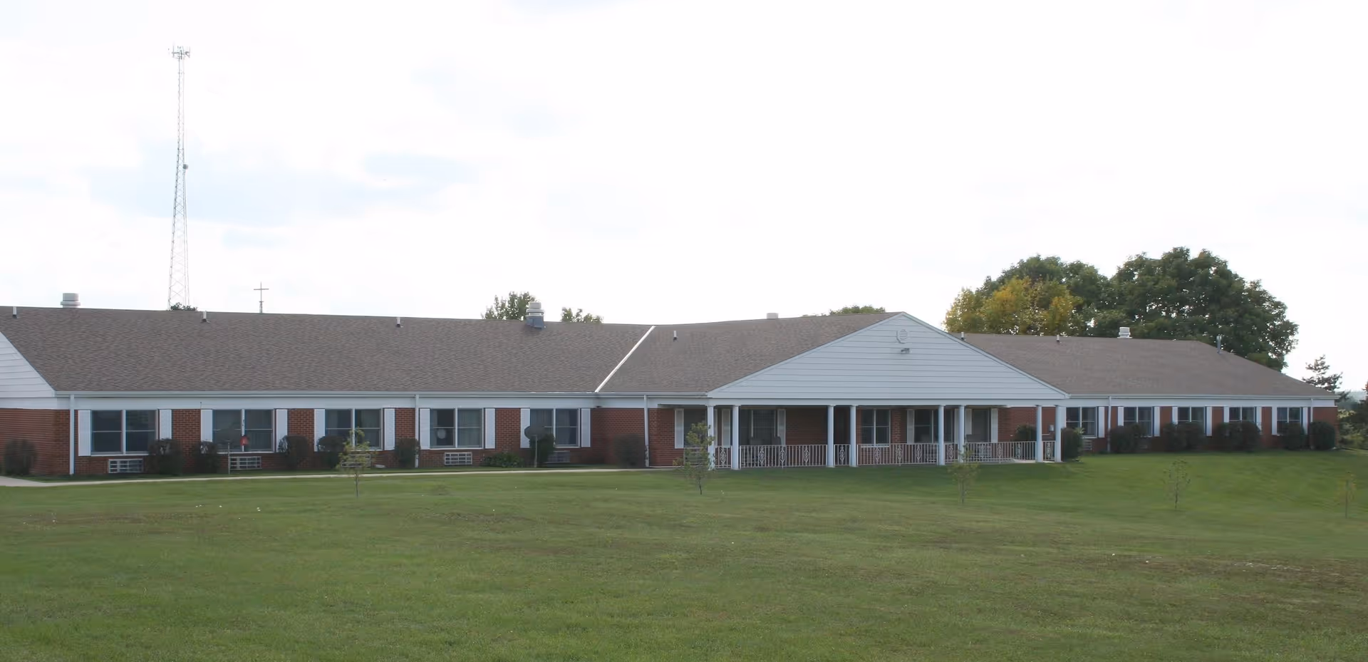 Exterior front view of a single-story brick building with a gray shingled roof, white trim, and a covered porch area. The building is surrounded by a large grassy lawn with a few small trees and bushes. The sky is partly cloudy.