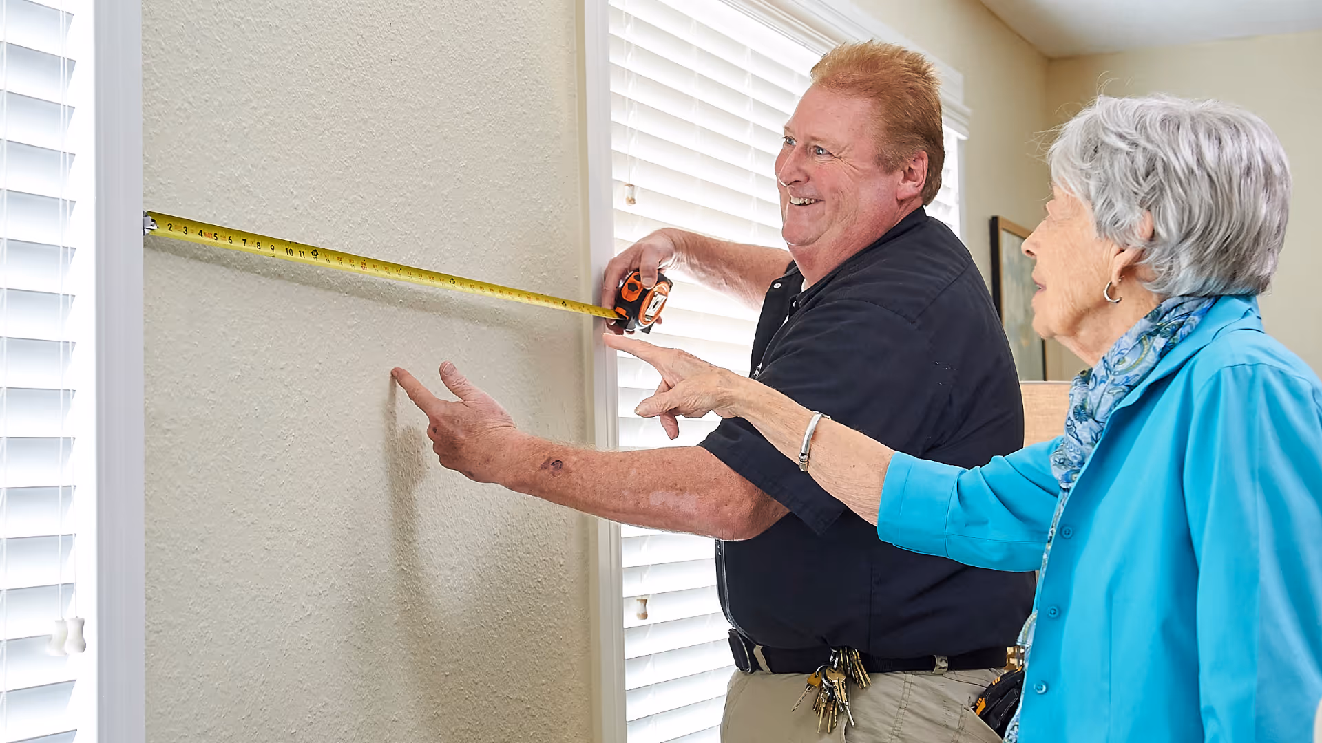 A man and an elderly woman are measuring the width of a wall between two windows with white blinds using a yellow tape measure. The man is holding the tape measure while the woman points at the wall. They are indoors in a well-lit room.