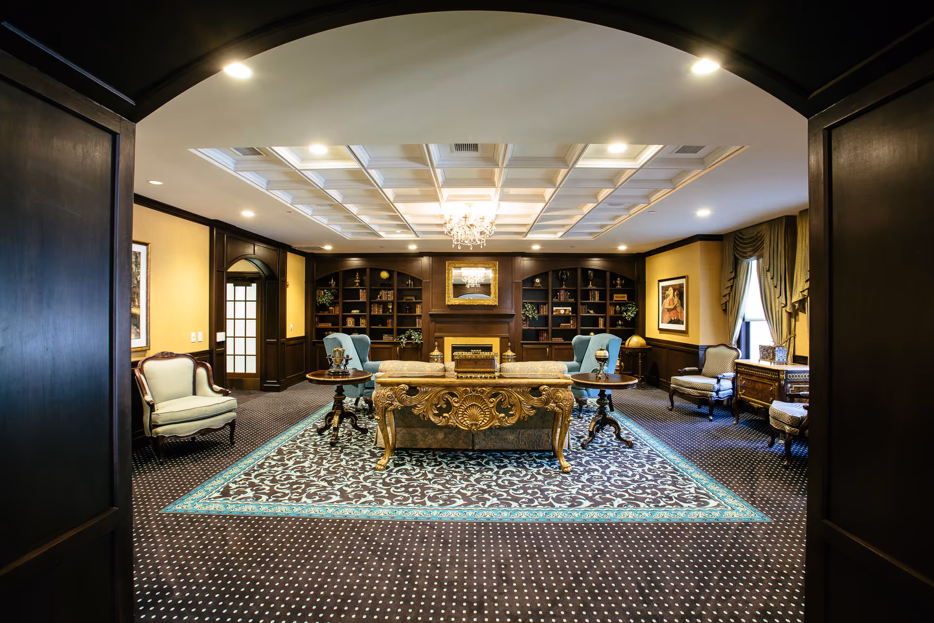 Ornately furnished lounge with a carved central table, armchairs, patterned rug and built-in bookcases under a coffered ceiling.