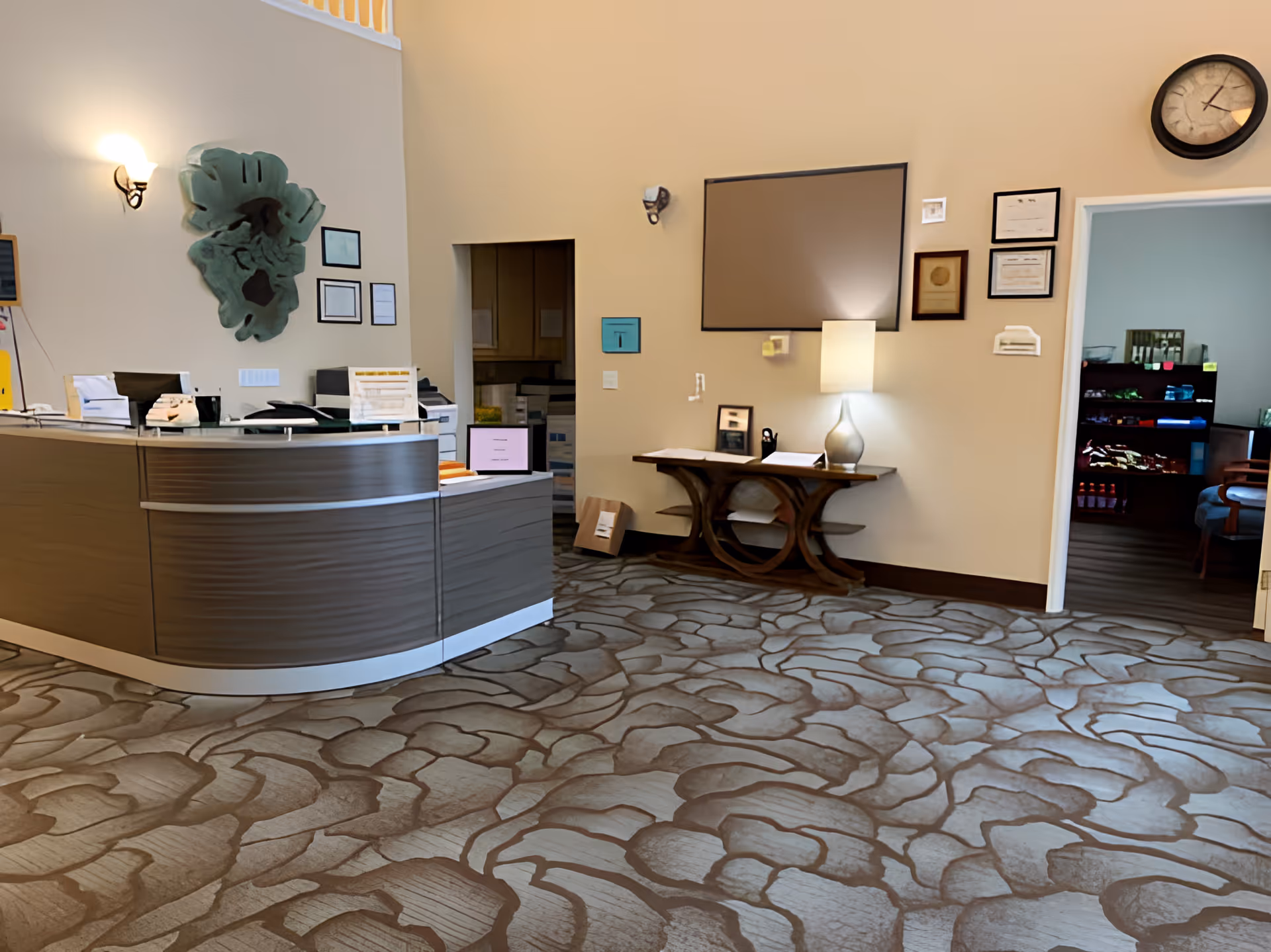 Reception area of a senior living facility with a curved wooden front desk, decorative wall art, a small table with a lamp, framed certificates on the wall, and a clock. An open doorway reveals a room with shelves stocked with various items.