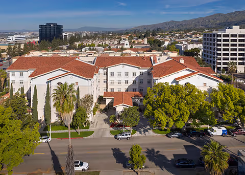 Aerial view of Belmont Village Senior Living Burbank showing a large white building with red-tiled roofs surrounded by trees and parked cars along the street. The background includes other buildings and distant mountains under a clear blue sky.