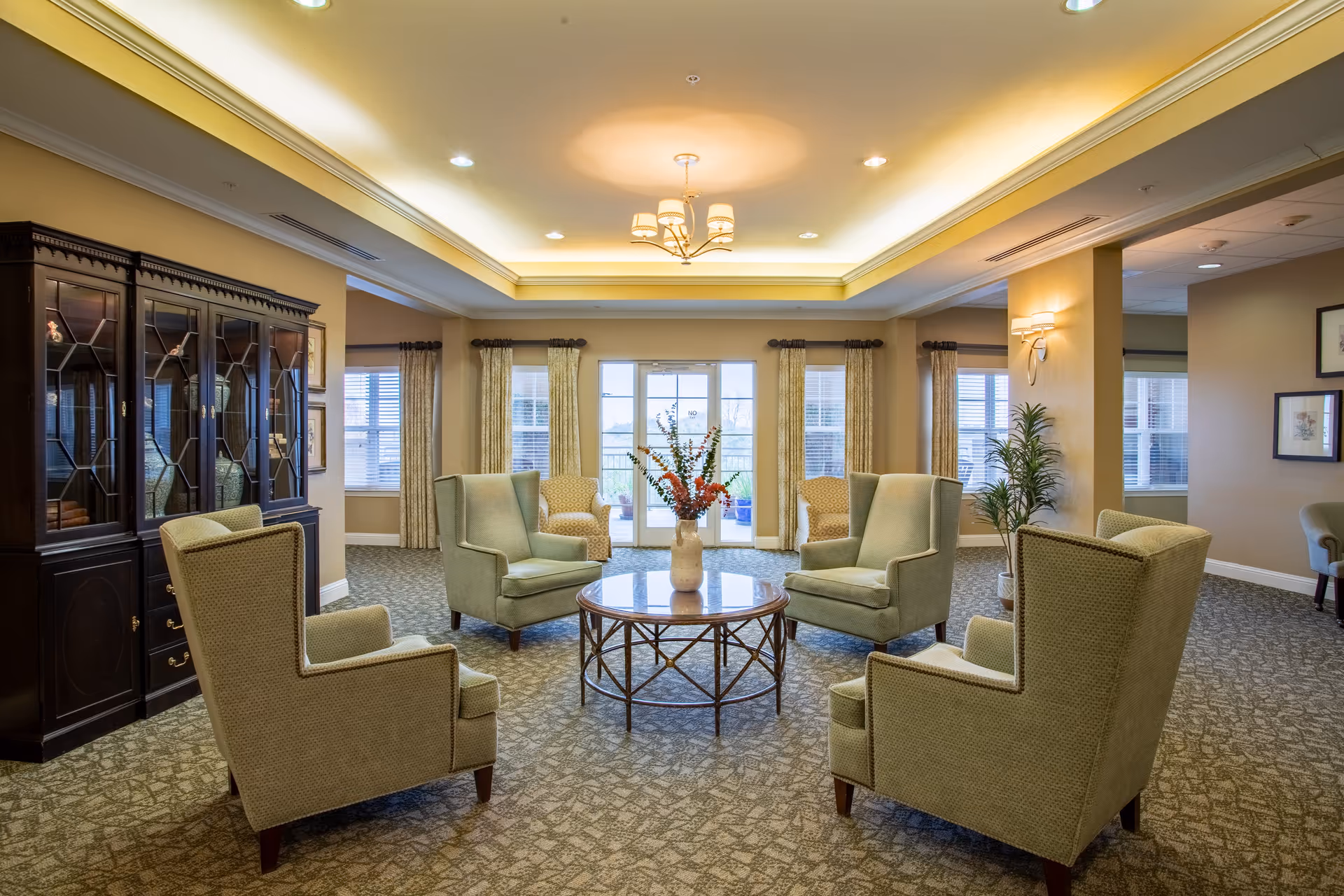 A cozy living room area in The Village of Meyerland featuring four green upholstered armchairs arranged around a round glass-top coffee table with a vase of flowers. The room has large windows with curtains, a decorative ceiling light fixture, a dark wooden cabinet on the left, and a potted plant near a column on the right.