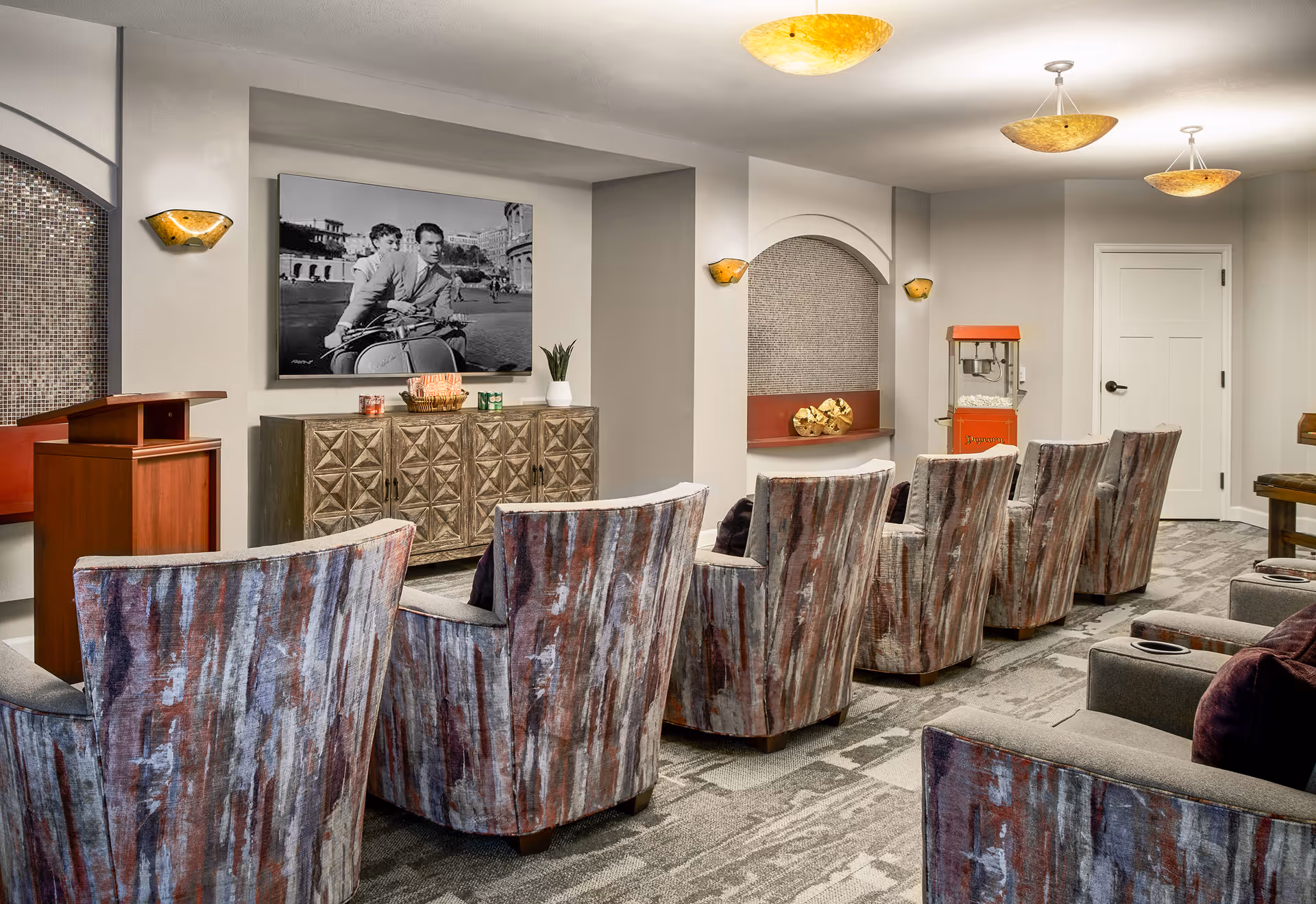 A cozy theater-style room with two rows of patterned armchairs facing a wall with a vintage black and white photo of a couple on a scooter. The room features warm lighting with ceiling and wall sconces, a wooden podium, a popcorn machine, and decorative elements including a cabinet and a small plant.