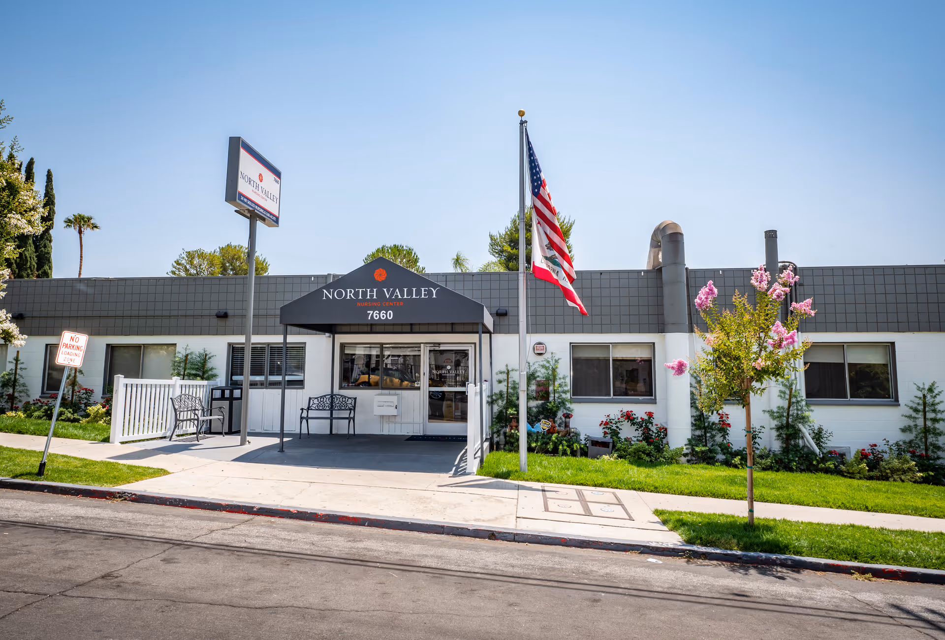 Front entrance of North Valley Nursing Center with a canopy reading 'North Valley 7660', an American flag, benches, and landscaped lawn.