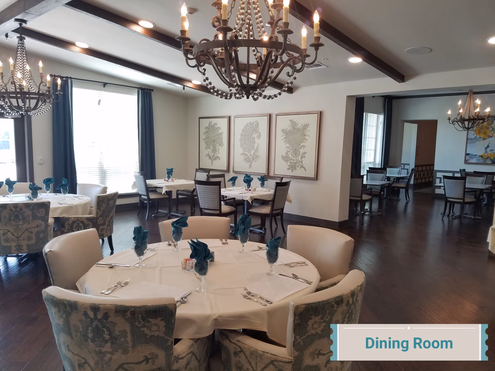 A well-lit dining room with multiple round and square tables covered with white tablecloths. Each table is set with silverware and glasses with teal-colored napkins folded inside. The room features dark wooden floors, large windows with dark curtains, decorative wall art, and ornate chandeliers hanging from the ceiling.