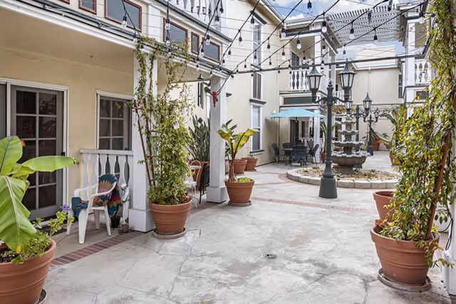Sunlit courtyard with potted plants, string lights, balconies, seating, and a central fountain.