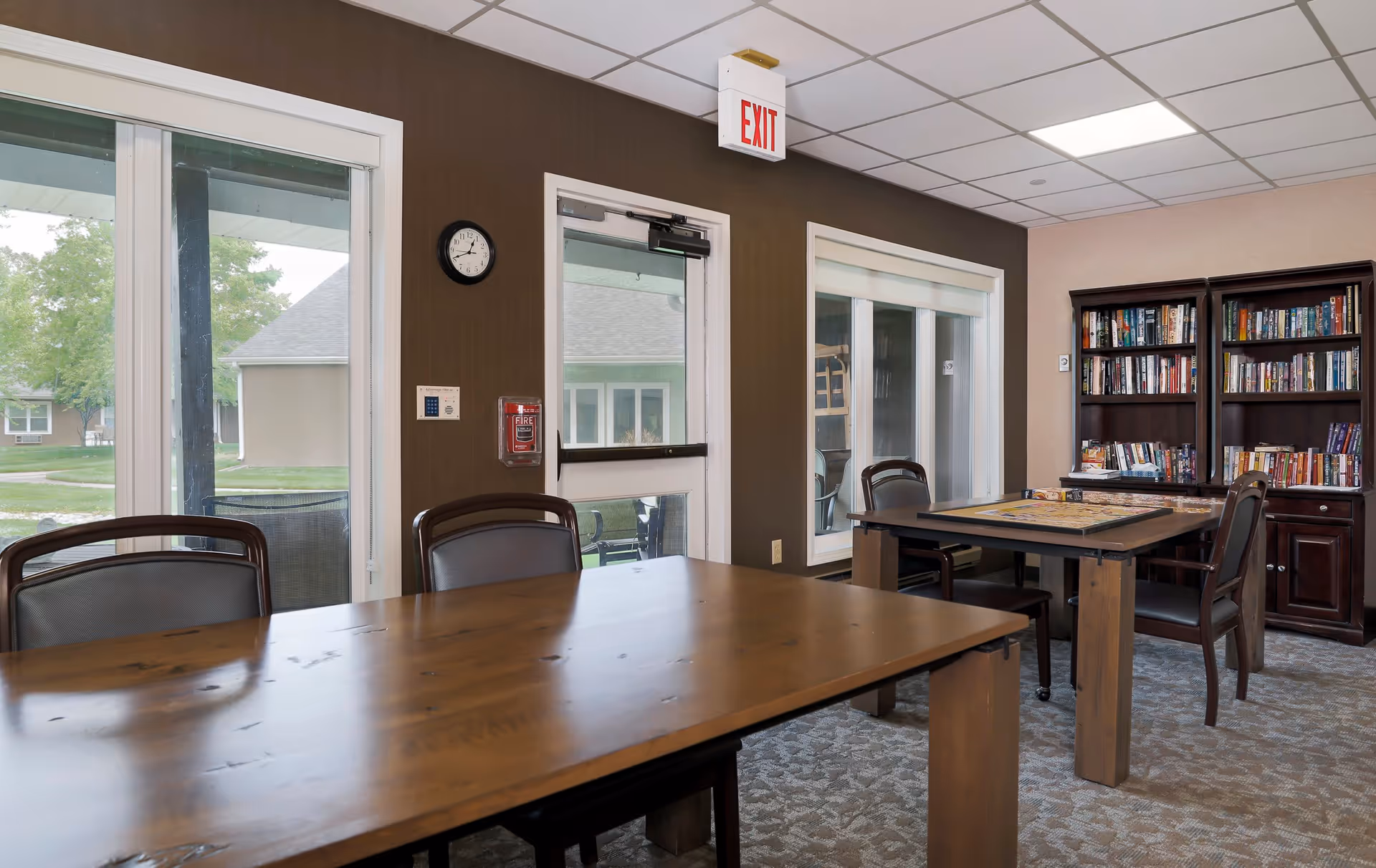A cozy interior room with two wooden tables and several chairs. One table has a board game set up on it. There are large windows and a glass door letting in natural light, with a view of green outdoor space. A clock, fire alarm, and exit sign are mounted on the wall. Two bookshelves filled with books are against the far wall.