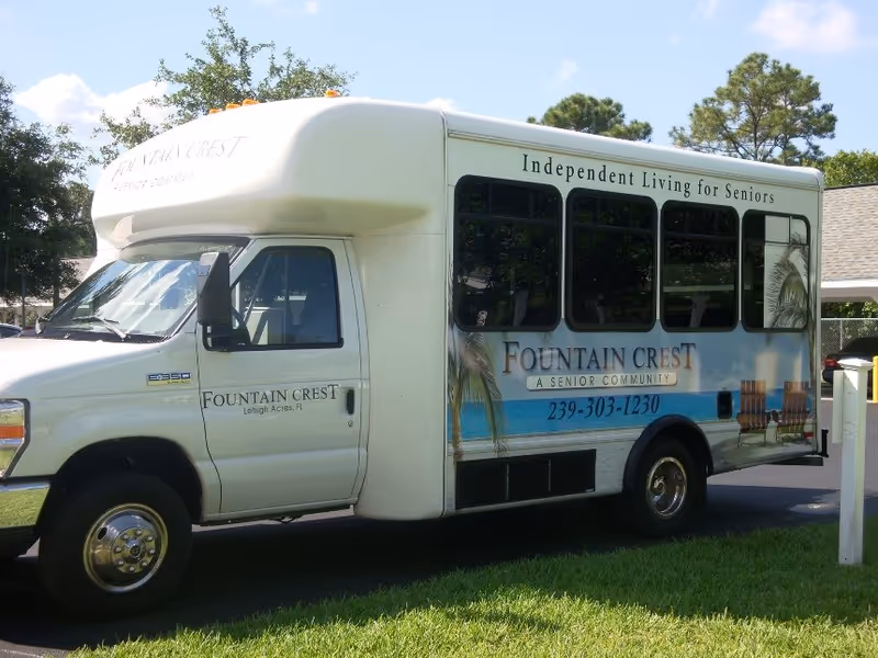 A white shuttle bus parked on grass near a driveway. The bus has signage for Fountain Crest Retirement Community, advertising independent living for seniors with a phone number. Trees and a building are visible in the background.