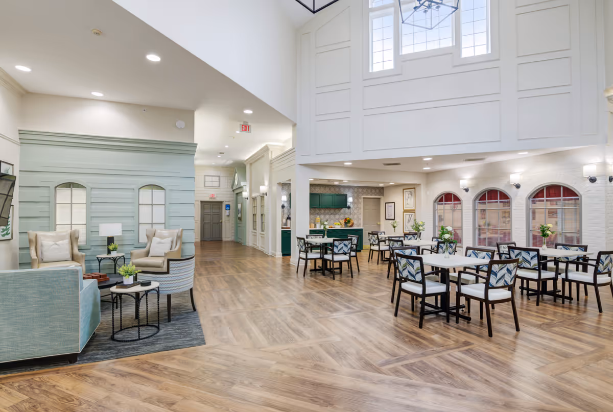 A spacious senior living community common area with a seating lounge on the left featuring armchairs and a sofa around small tables, and a dining area on the right with multiple tables and chairs. The room has high ceilings with large windows, wood flooring, and soft lighting.
