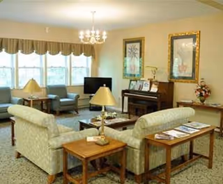 A cozy living room area in a senior living facility with two patterned sofas arranged around a coffee table, a piano against the wall, framed artwork, a television, and several lamps providing warm lighting.