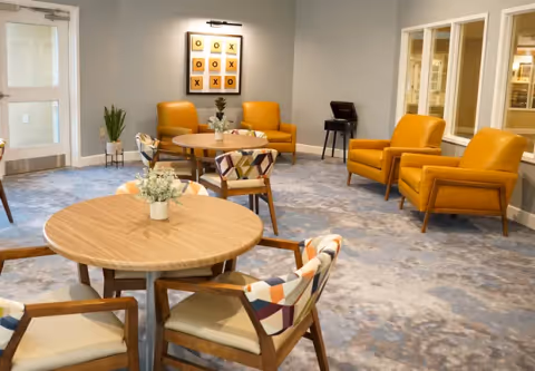 A bright communal seating and dining area with round wooden tables, patterned chairs, and mustard-yellow armchairs in a memory care facility.