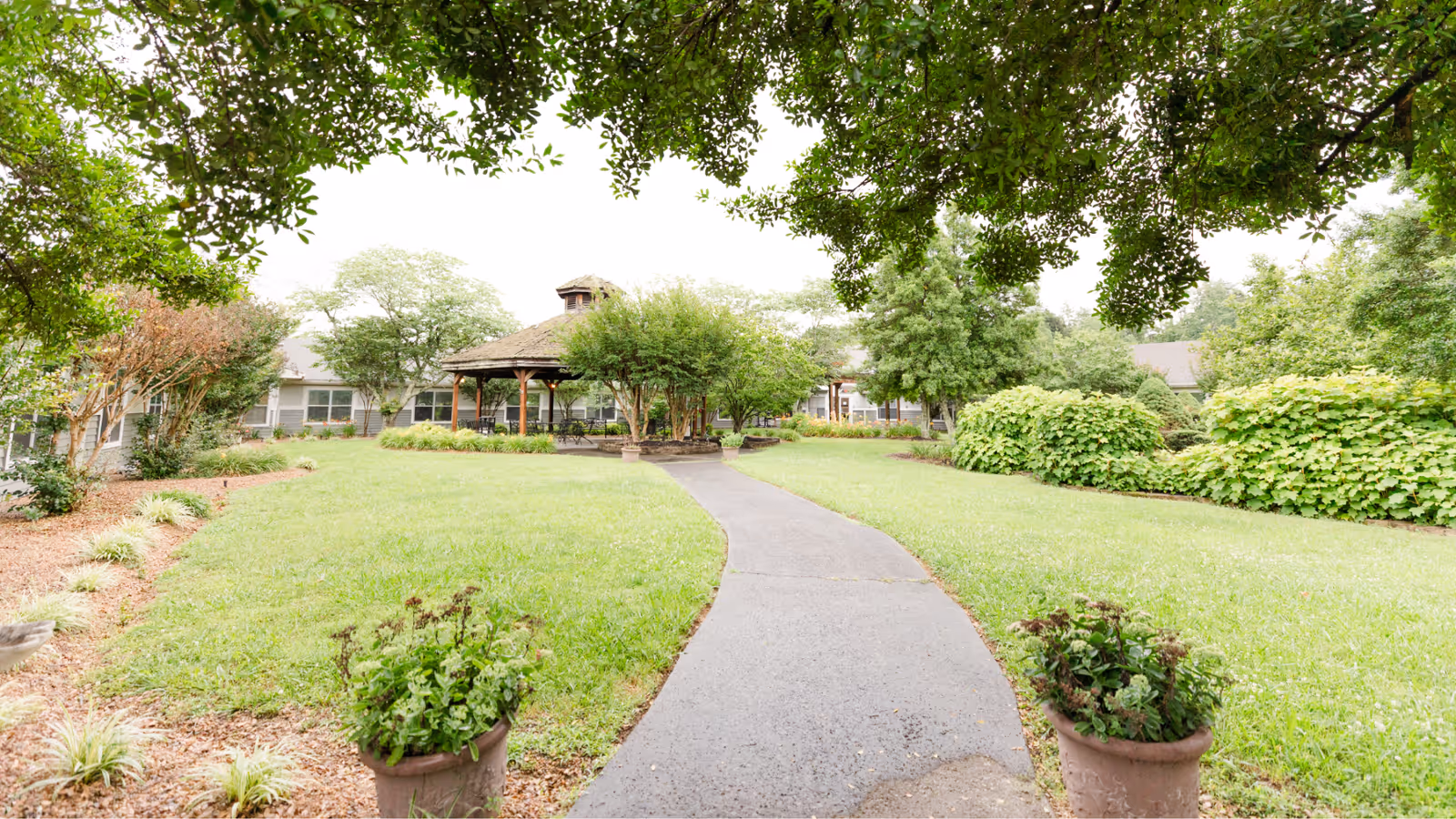 A paved walkway leads through a well-maintained garden with green grass, shrubs, and trees towards a gazebo in front of a single-story building. The scene is framed by tree branches overhead.
