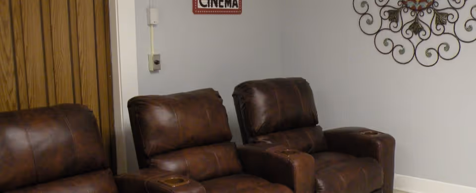 A cozy room with three brown leather recliner chairs arranged side by side. On the wall above the chairs, there is a decorative metal wall art and a small sign that reads 'CINEMA'. The room has a wooden door and light-colored walls.