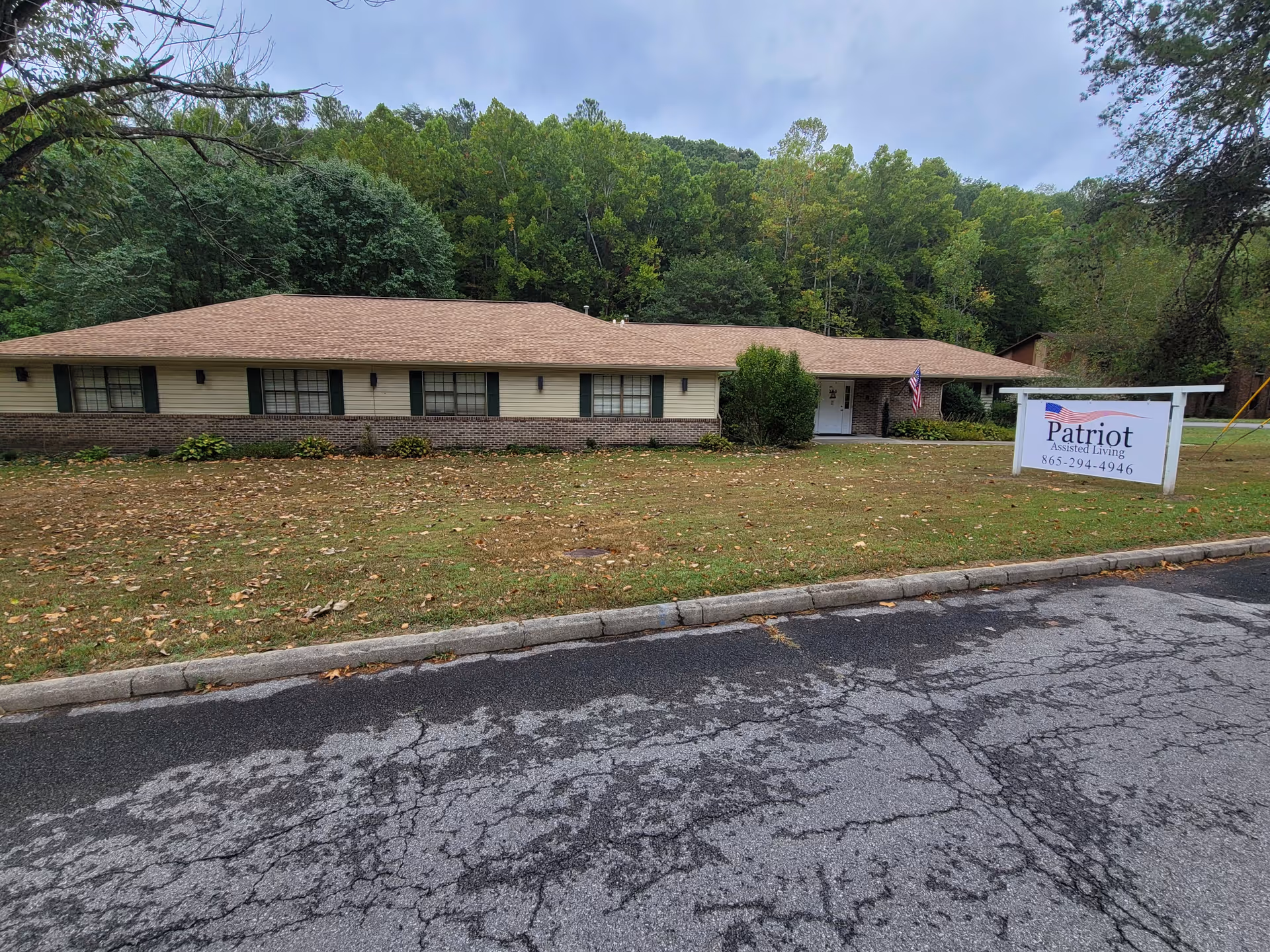 Single-story brick-and-siding assisted living building with a front lawn, American flag, and a sign reading 'Patriot Assisted Living'.
