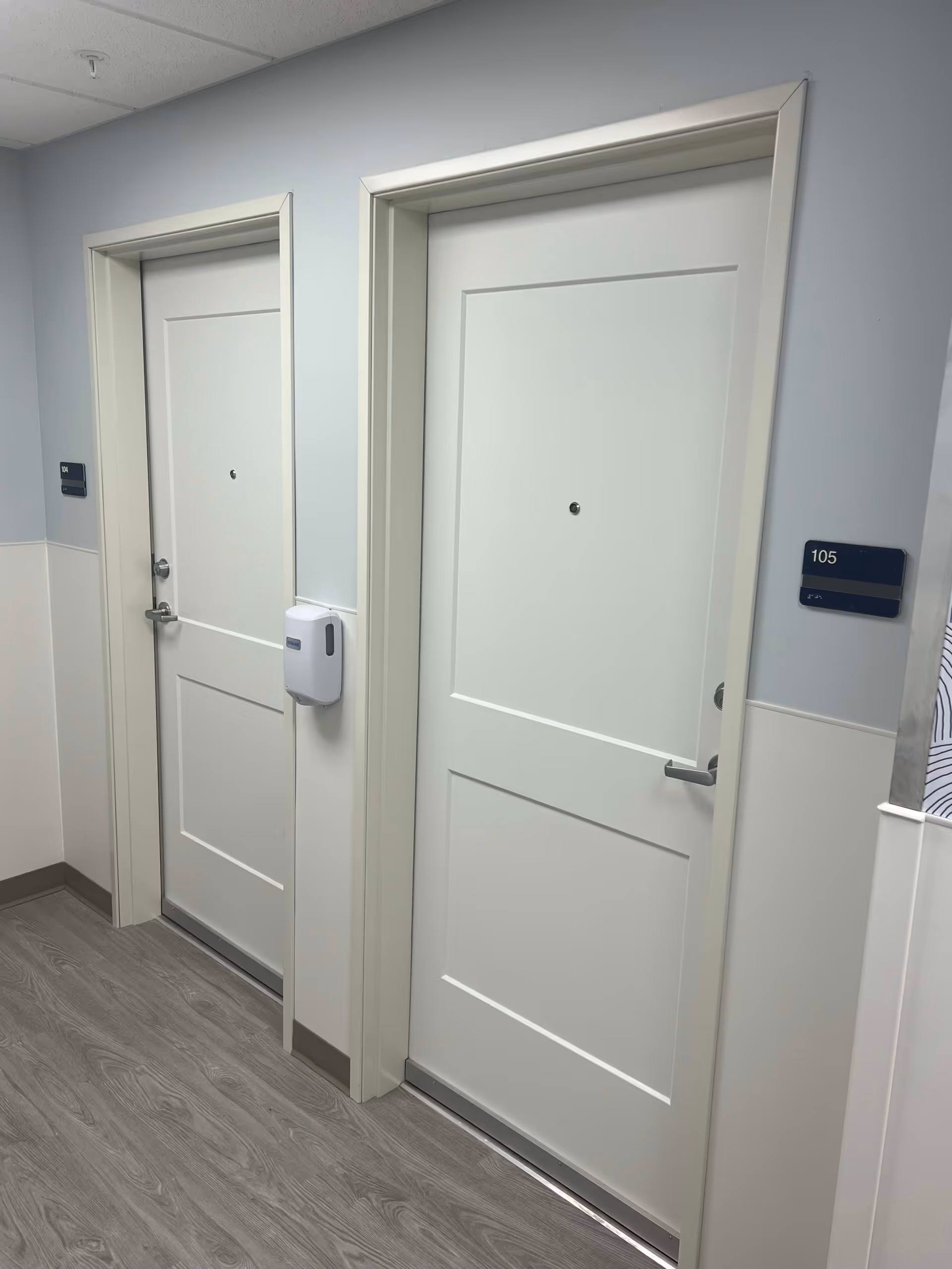 Interior hallway with two white apartment doors, one labeled 105, and a wall-mounted sanitizer dispenser between them.