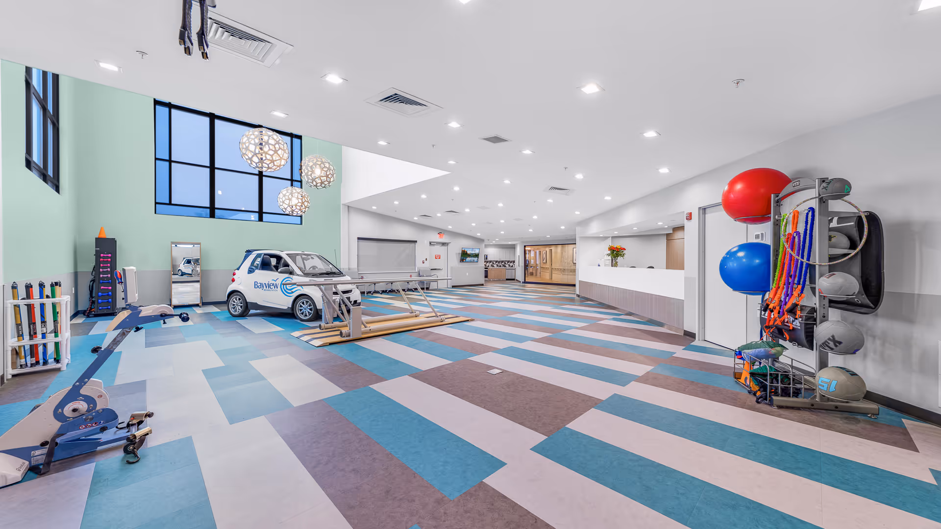 Spacious, brightly lit rehab interior with a colorful patterned floor, exercise equipment (including therapy balls and a small branded car) and a reception desk.