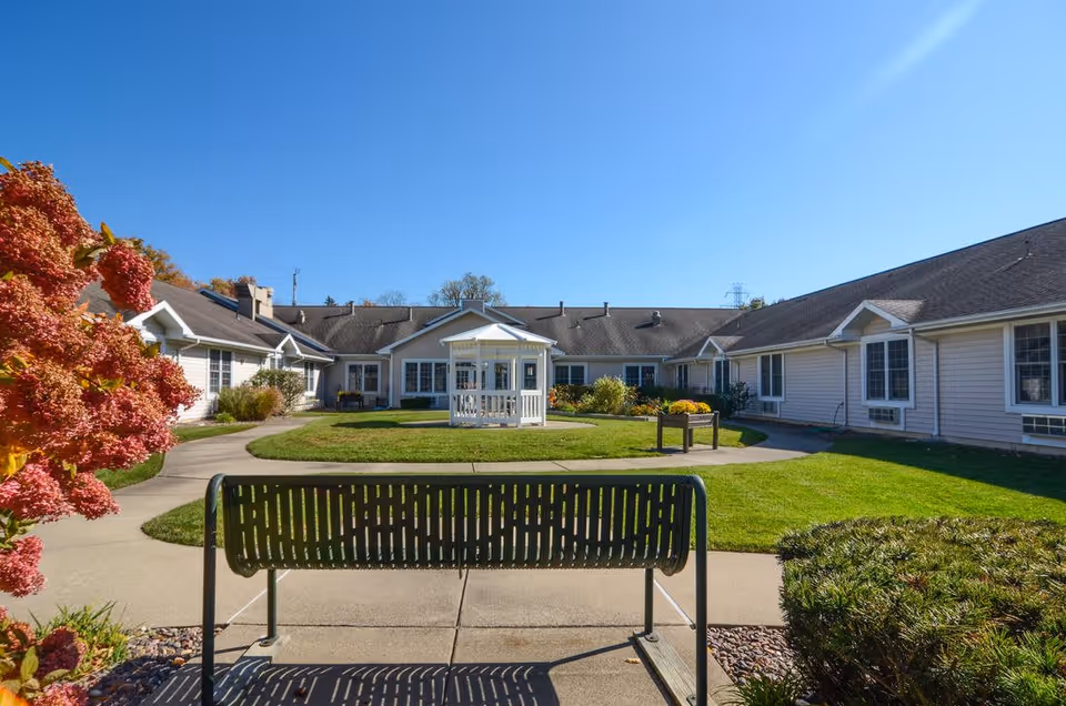 Outdoor courtyard area of a senior living community with a green metal bench in the foreground, a paved walkway, a white gazebo in the center, surrounded by well-maintained grass and shrubs, and single-story buildings with beige siding under a clear blue sky.