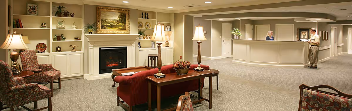 A cozy nursing center common area with a red sofa, two floral armchairs, a wooden table with two lamps, built-in white shelves with decorative items, and a fireplace. In the background, a reception desk with a woman standing behind it and a man talking to her. The walls are painted beige and the floor is carpeted.