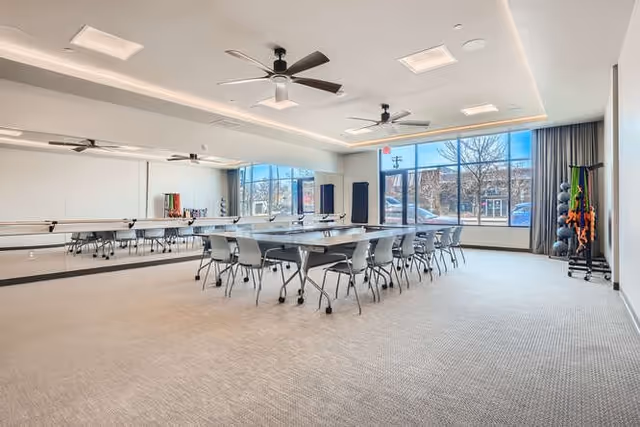 A spacious, well-lit room with large windows showing an outdoor view. The room contains several tables and chairs arranged in a rectangular formation, ceiling fans, and exercise equipment such as weights and resistance bands in the corner. The walls are light-colored, and one wall features a large mirror.