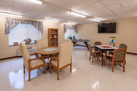 A well-lit common room with two tables surrounded by chairs, a foosball table against the far wall, a bookshelf filled with books, two windows with floral curtains, and a wall-mounted flat screen TV.