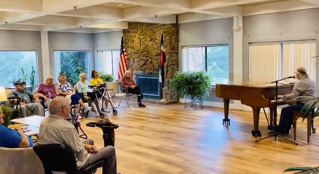 A group of elderly people seated in a spacious room with large windows, listening to a man playing a grand piano and singing into a microphone. The room has wooden floors, a stone fireplace with American and Colorado state flags, and several potted plants.