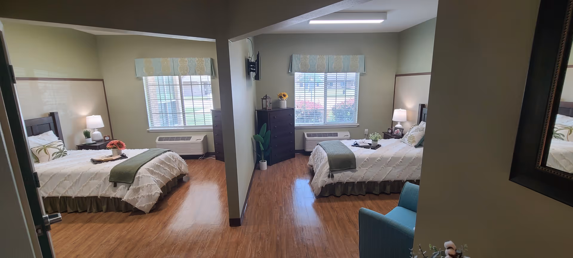 A bright and clean shared bedroom in a senior living facility with two beds separated by a partial wall. Each bed is neatly made with white bedding and green accents. The room features wooden flooring, two windows with blinds and valances, bedside tables with lamps, a dresser, and a blue armchair. The overall decor is simple and welcoming.