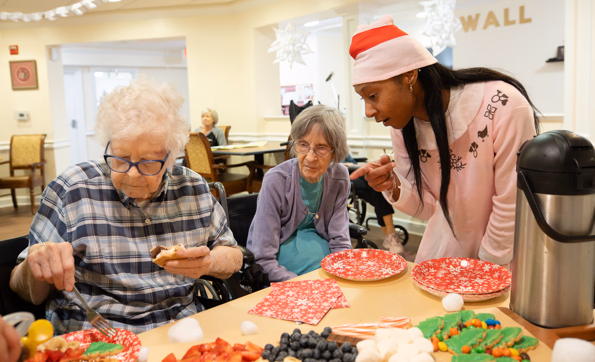 Two elderly women sitting at a table with holiday-themed plates and snacks, while a younger woman wearing a Santa hat leans over to talk to them in a festive indoor setting.