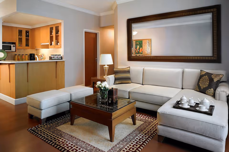 Contemporary living room with a light sectional sofa, ottoman, coffee table with flowers, and an open kitchen in the background.